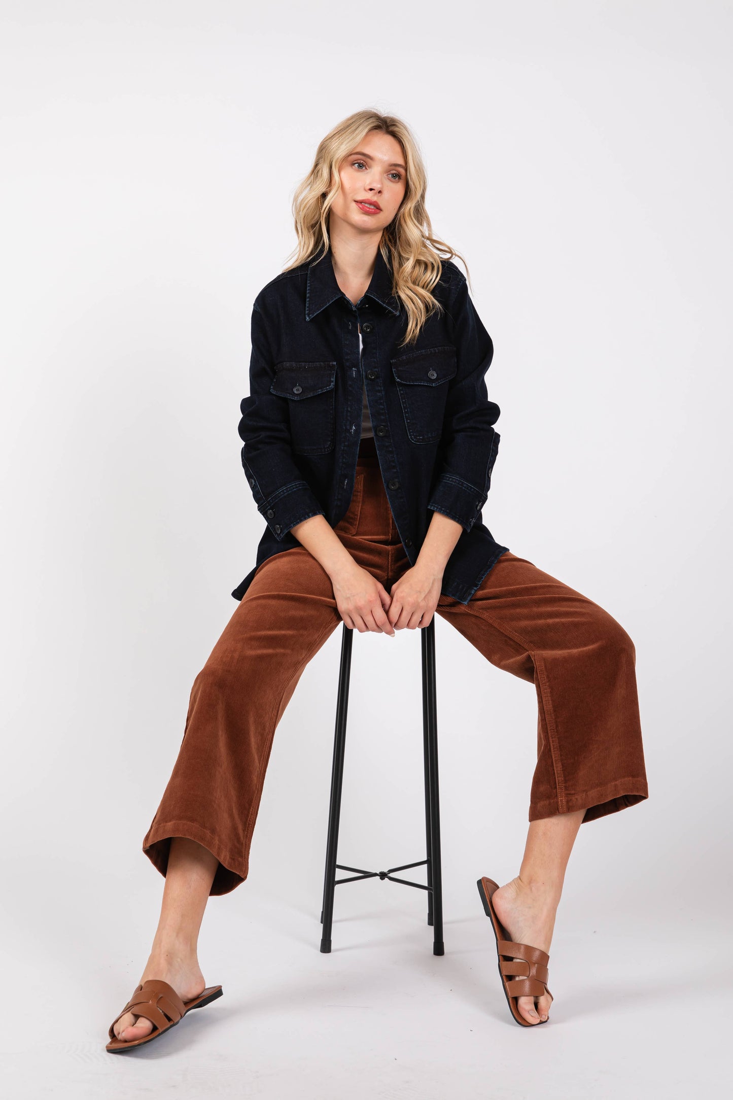Woman sitting on a stool wearing a black jacket and brown pants against a white background