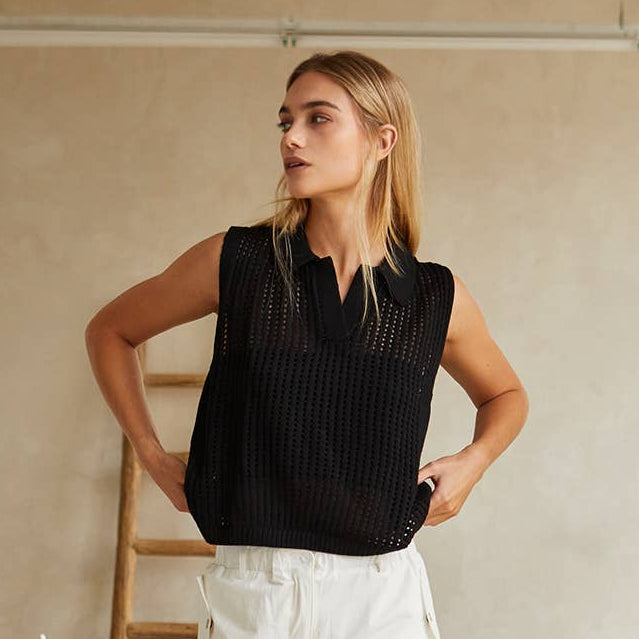Woman wearing a black sleeveless top and white pants in a minimalistic room.