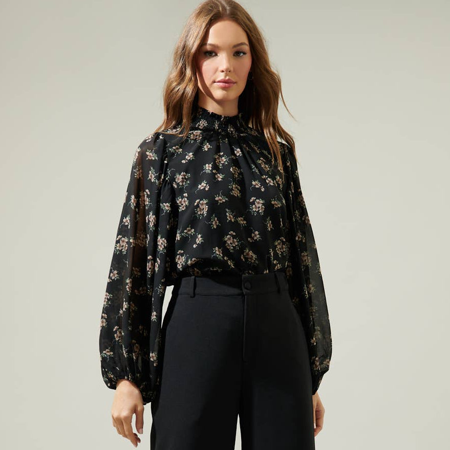 
                      
                        Woman wearing a black floral blouse and black pants on a plain background
                      
                    