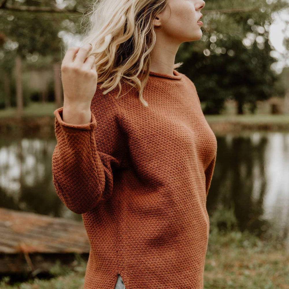 Woman wearing a rust-colored sweater standing outdoors with trees and water in the background