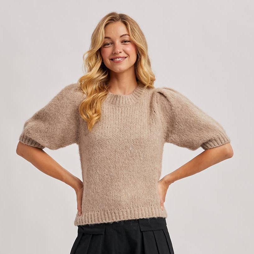 Woman wearing a beige sweater and black pants on a plain background