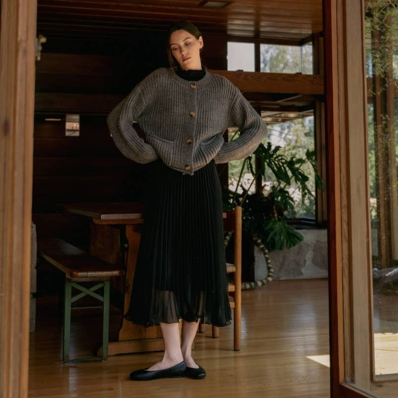 
                      
                        Woman wearing a gray knitted cardigan and black pleated skirt standing in a room with wooden floors and furniture.
                      
                    