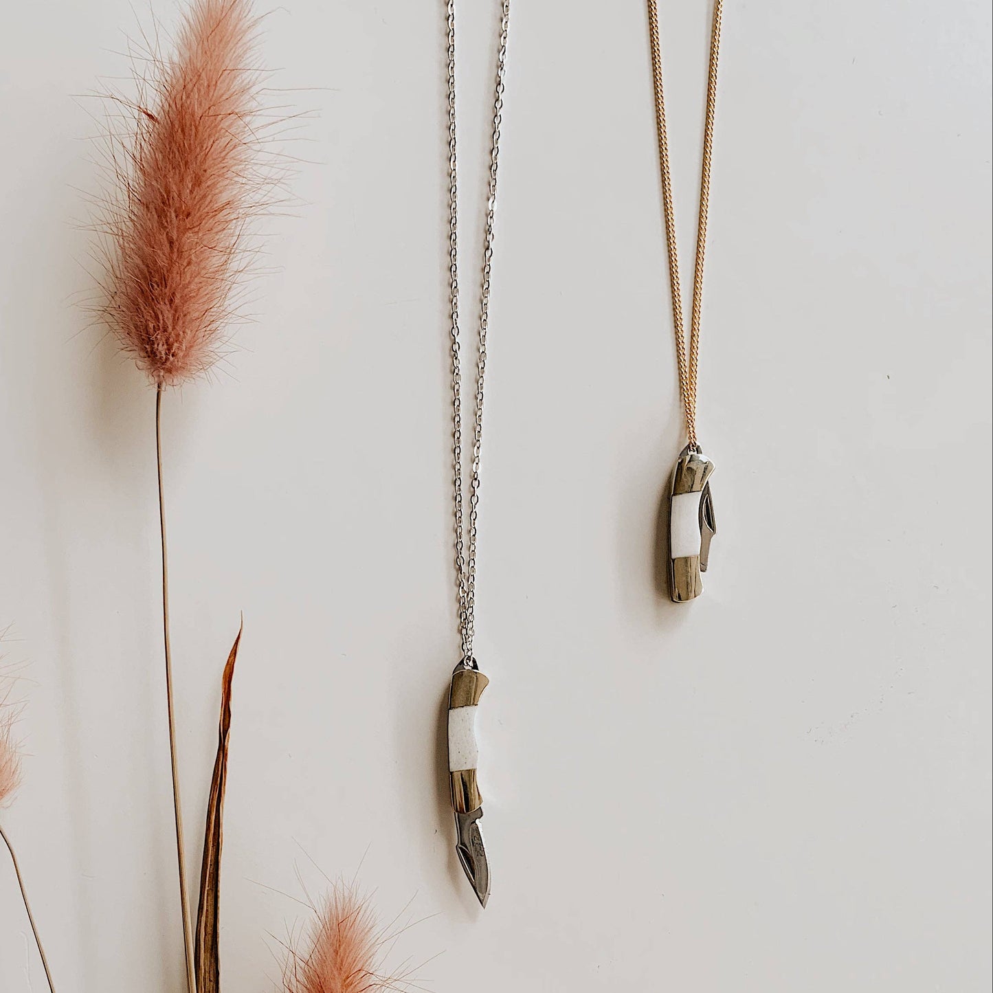 Two necklaces hanging on a white wall with pink dried grasses.
