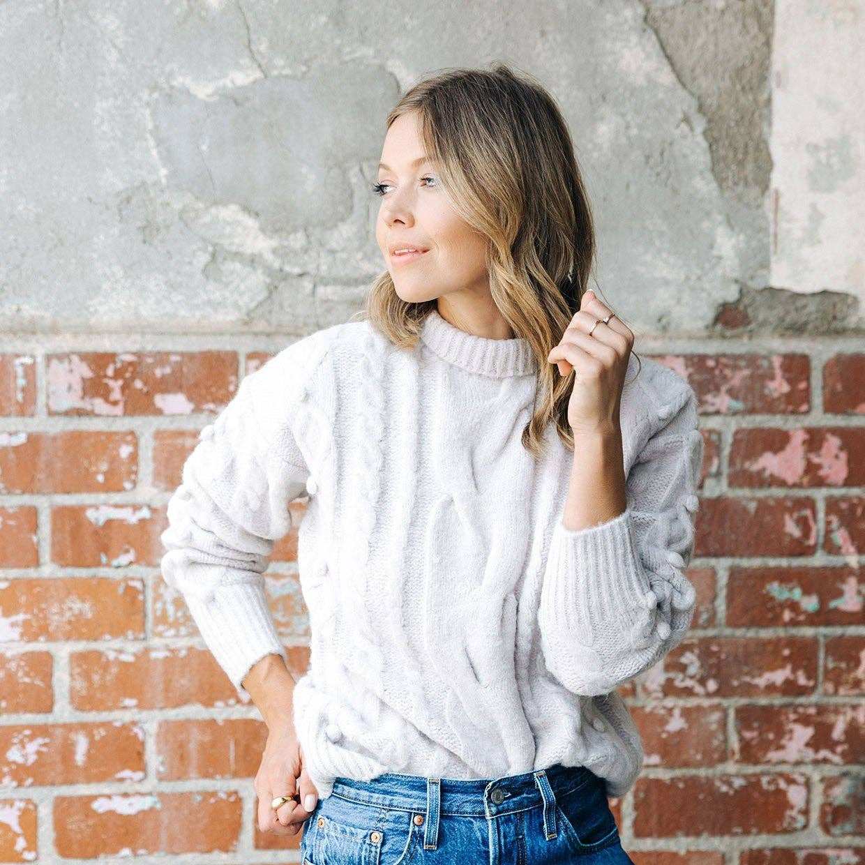 Woman wearing a white sweater and denim shorts standing against a brick wall.