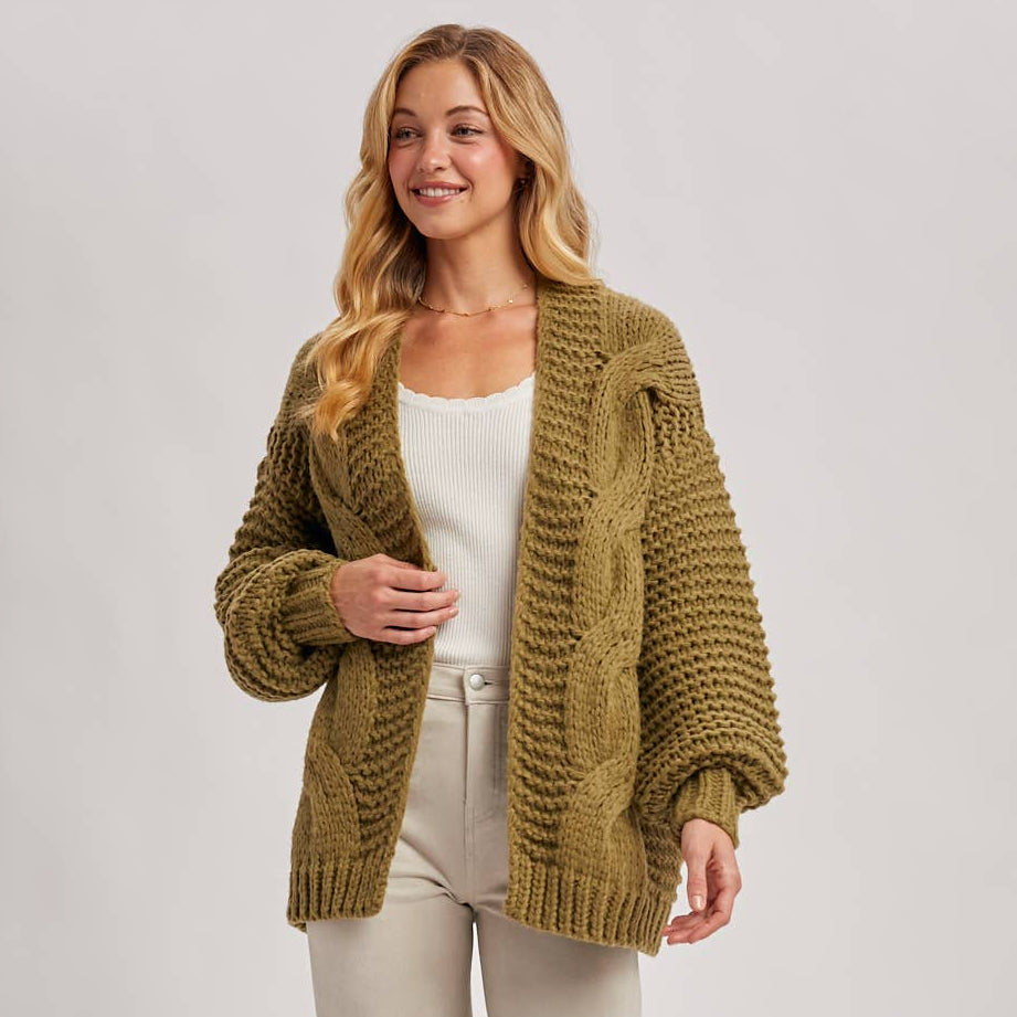 Woman wearing a green knitted cardigan over a white top and beige pants on a light gray background